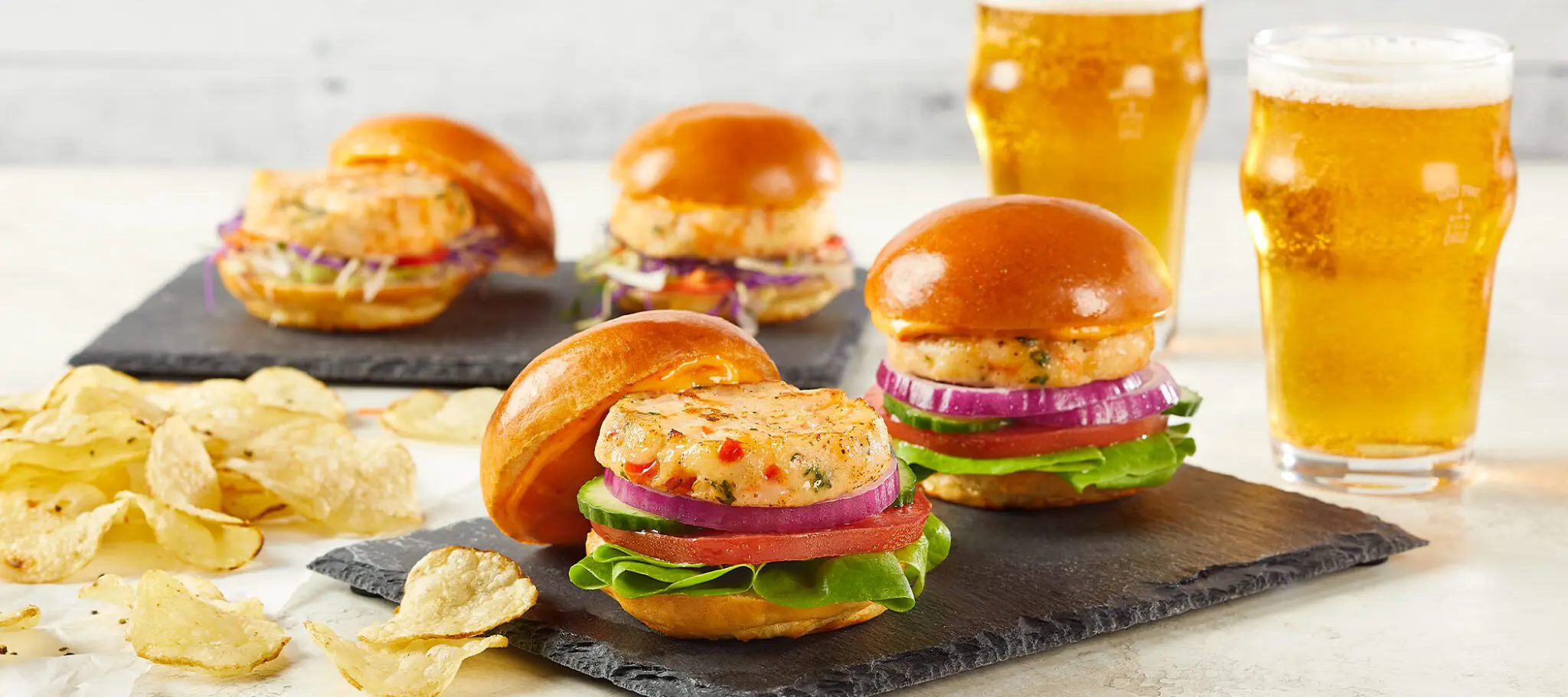 Social Kitchens Pro Premium Shrimp Sliders on a slate slab served with scattered potato chips and glasses of beer.