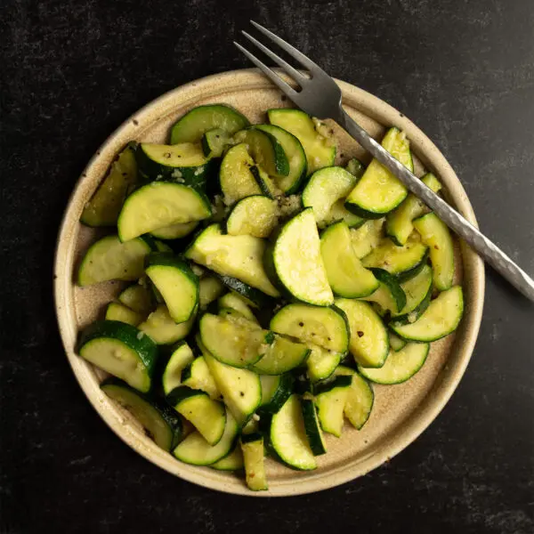 Overhead view of garlic zucchini on a beige plate over a textured black tabletop with a silver fork off to the side.