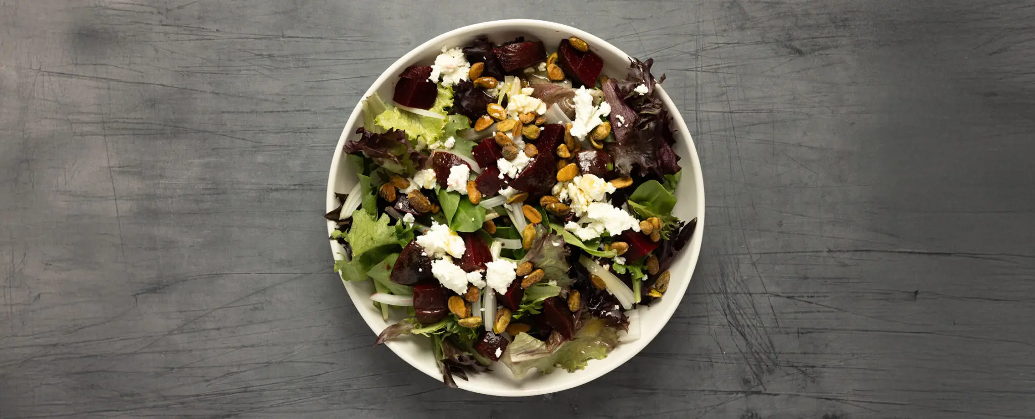 Overhead view of a Beet + Goat Cheese salad on a white plate served over a textured gray tabletop.