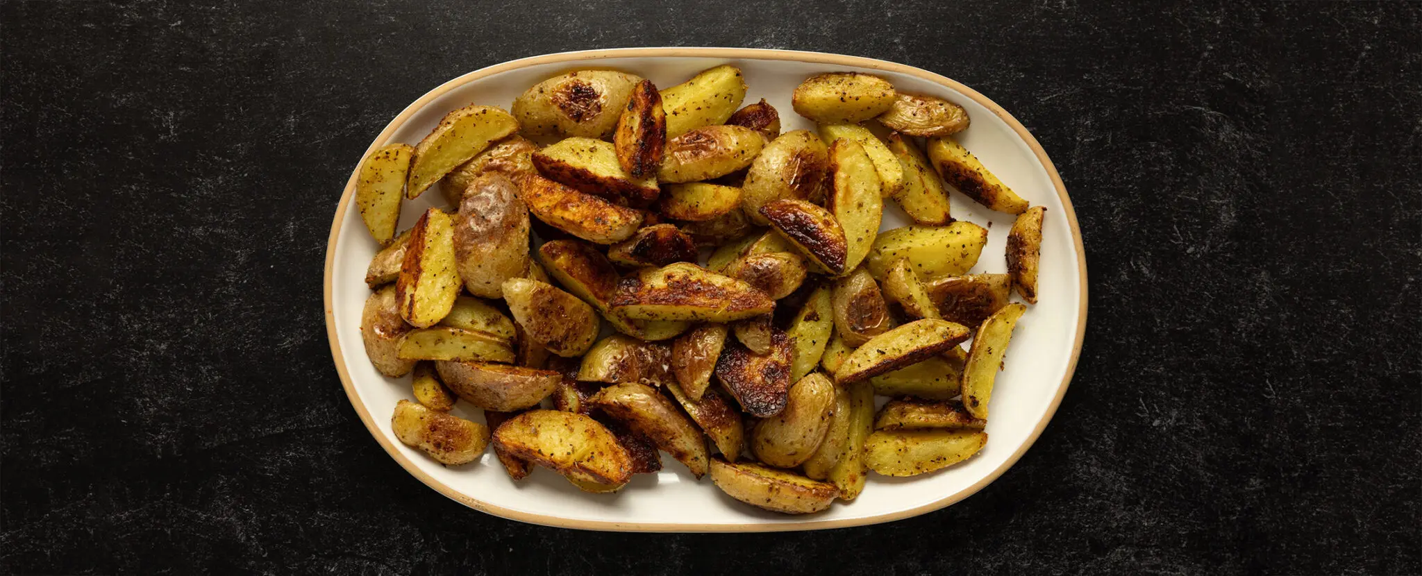 Overhead view of a wide white platter of Crispy Adobo Potato wedges served over a black stone tabletop.