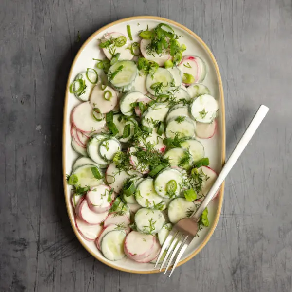 Overhead view of a large white platter of Creamy Cucumber & Radish Salad and a fork, served over a light gray textured tabletop.