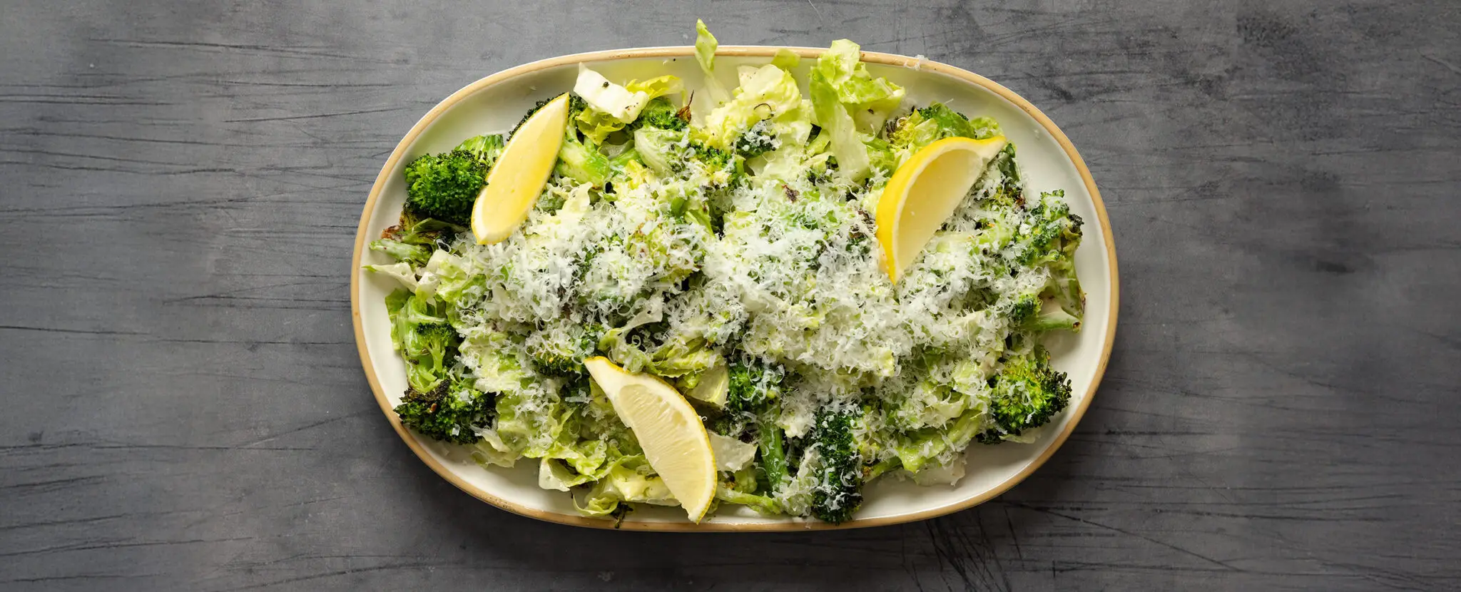 Overhead view of a large ceramic platter of Roasted Broccoli Caesar Salad with lemon wedges, served over a gray textured tabletop.