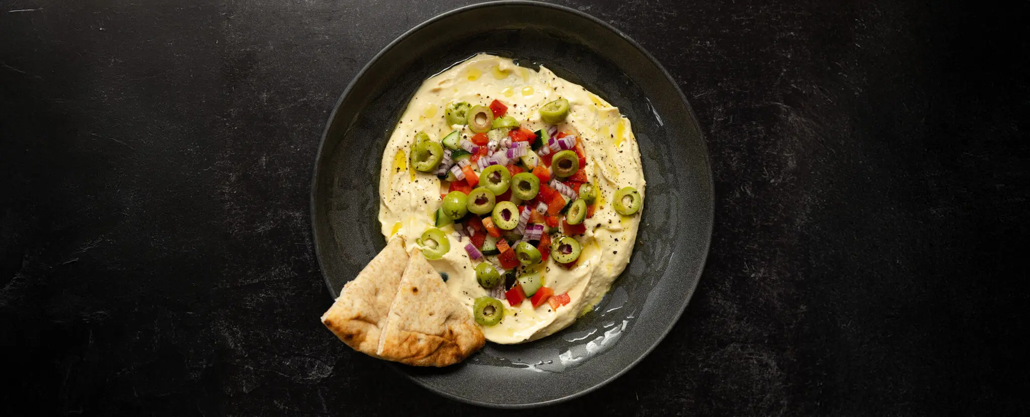 Overhead view of a black bowl of hummus topped with olives, red bell pepper, red onion, and cucumbers with a couple slices of pita bread.
