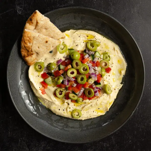Overhead view of a black bowl of hummus topped with olives, red bell pepper, red onion, and cucumbers with a couple slices of pita bread.