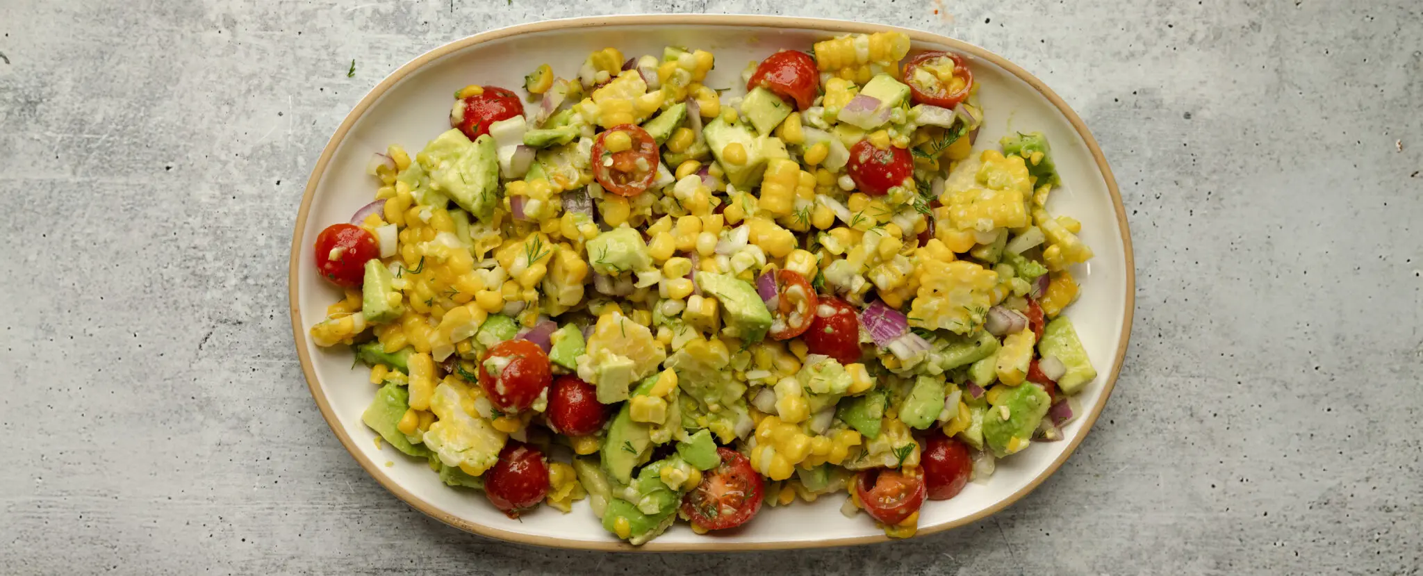 Overhead view of a large white ceramic plater of Corn & Avocado Salsa, served over a textured light gray stone tabletop.