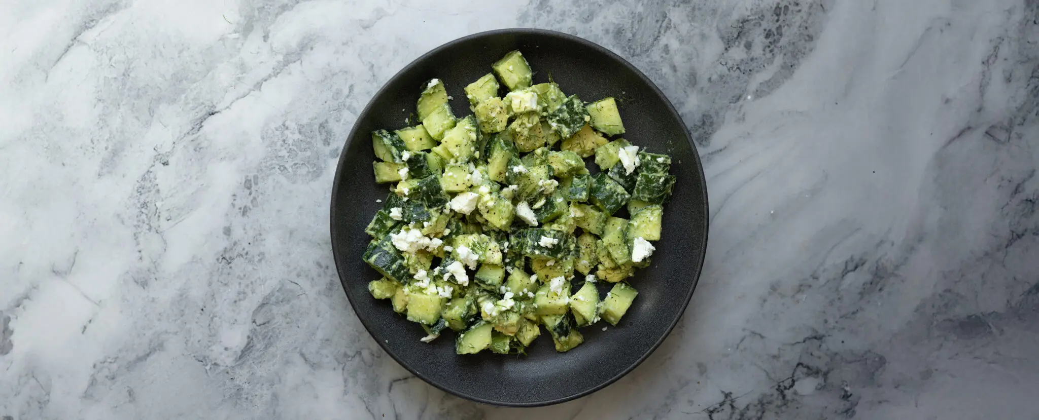 Overhead view of a cucumber feta salad served on a black plate over a marble tabletop.