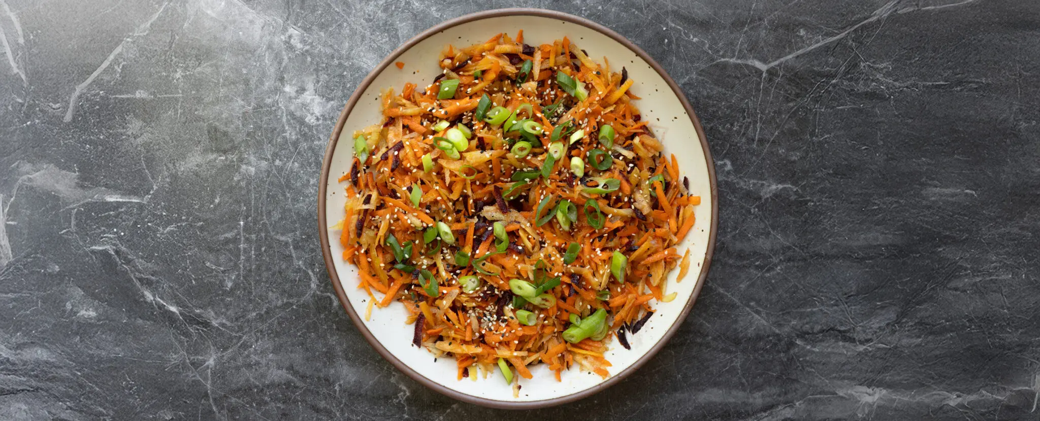 Overhead view of a bowl of sesame rainbow carrot salad garnished with green onions and served over a stone tabletop.