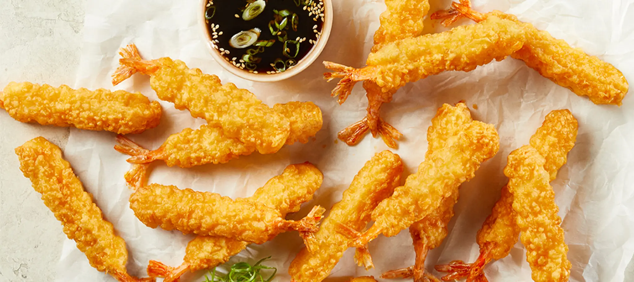 Overhead view of Social Kitchens Professional Tempura Battered Shrimp served over parchment paper with a small side of soy sauce with sesame seeds & scallions.