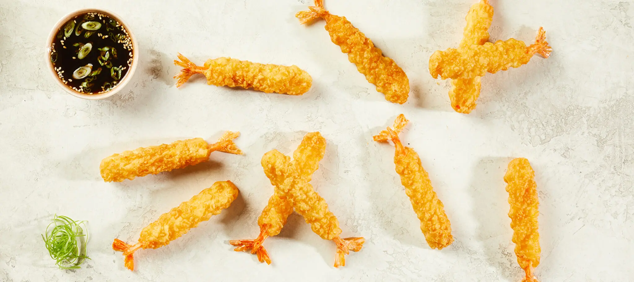 Overhead view of scattered pieces of Social Kitchens Professional Tempura Battered Shrimp served over parchment paper with a small side of soy sauce with sesame seeds & scallions.