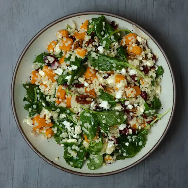 Overhead view of a white plate of Fall Couscous Salad topped with Social Kitchens Sweet & Spicy Shrimp, served on a gray wooden tabletop.
