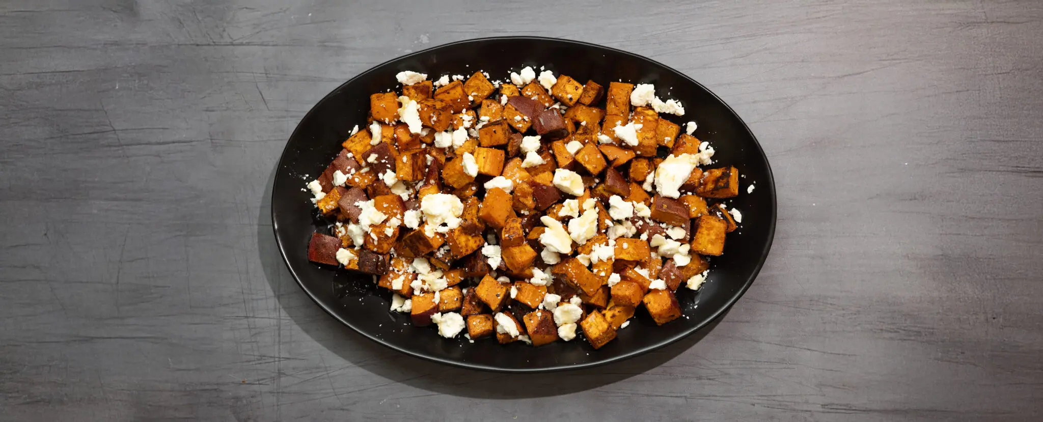 Three-quarter view of a large black platter of roasted chipotle sweet potatoes with crumbled feta on top, served over a dark gray wooden tabletop.