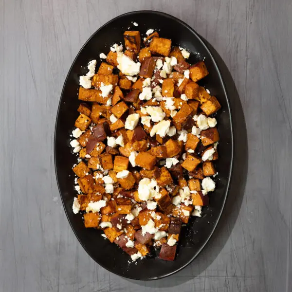 Overhead view of a large black platter of roasted chipotle sweet potatoes with crumbled feta on top, served over a dark gray wooden tabletop.