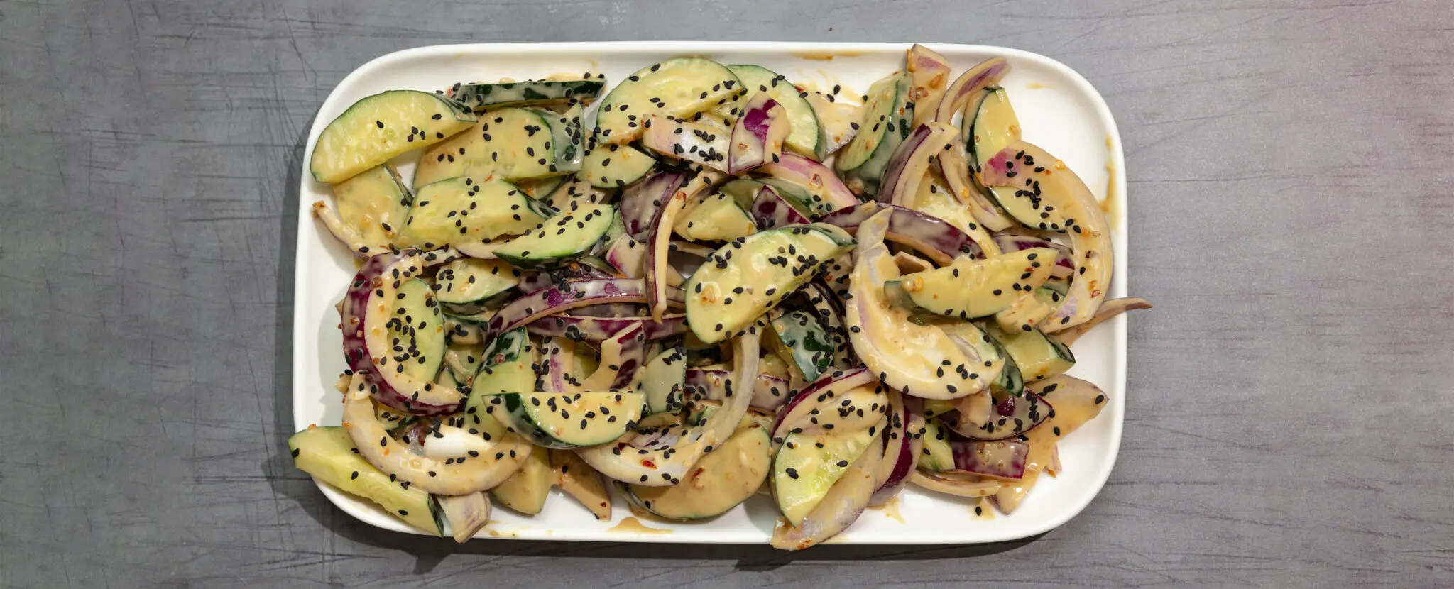 Overhead view of a large white rectangular plate of Tahini Cucumber Salad, served on a gray wooden tabletop.