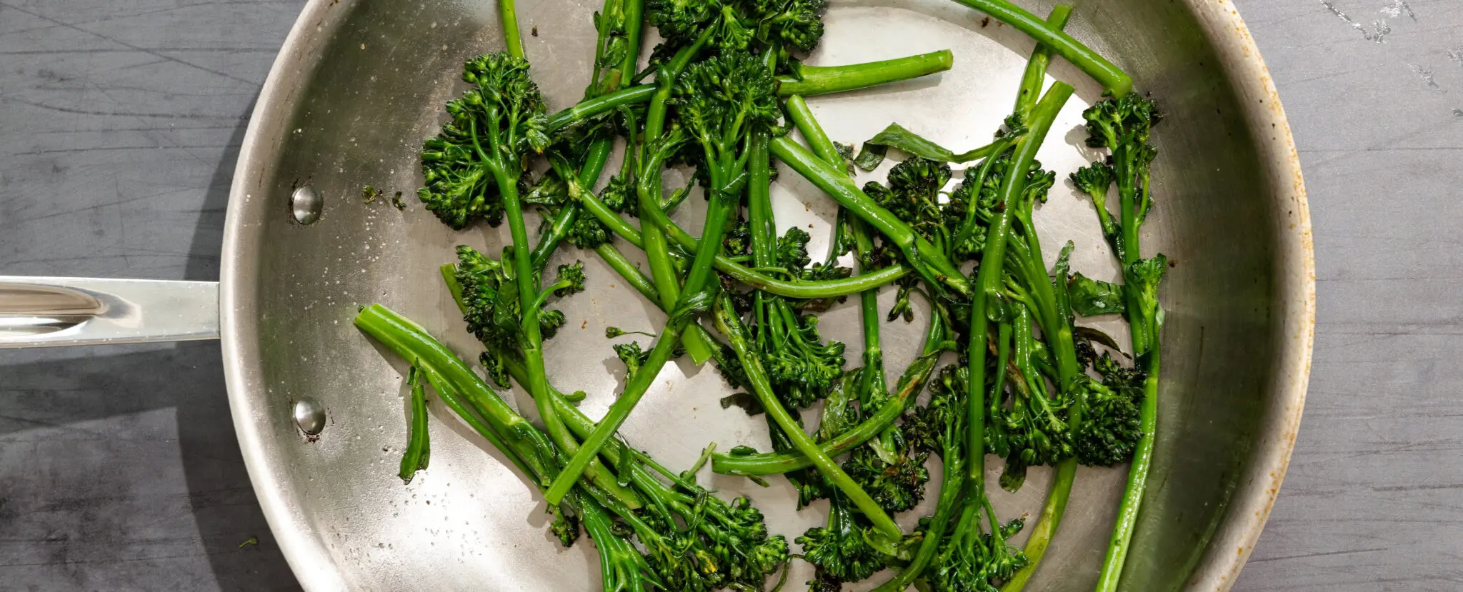 Overhead view of Simply Sautéed Broccolini in a stainless steel pan over a gray table surface.