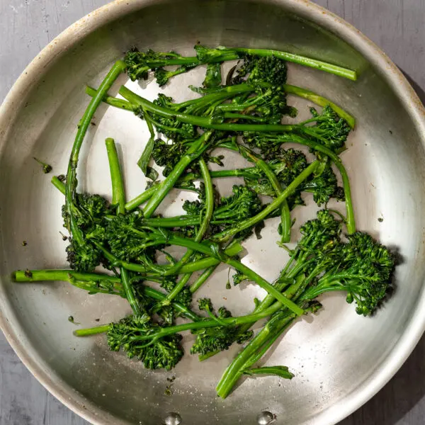 Overhead view of Simply Sautéed Broccolini in a stainless steel pan over a gray table surface.
