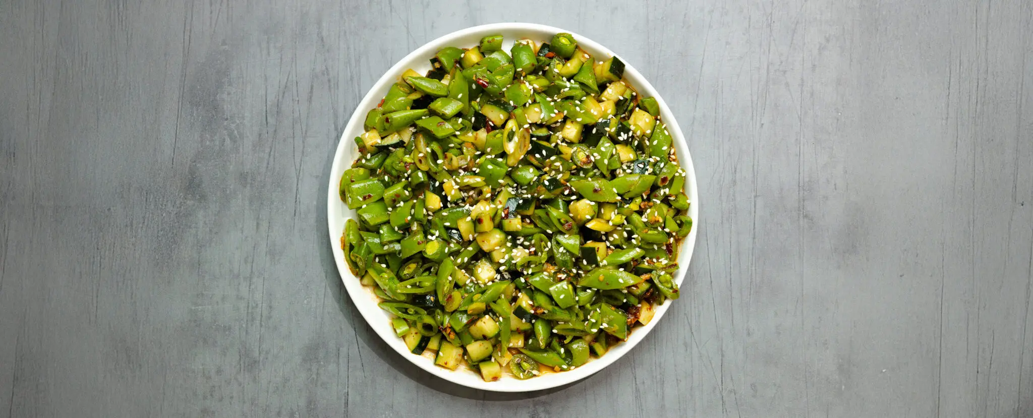 Overhead view of snap pea salad garnished with sesame seeds on a round white plate over a grey wooden tabletop.
