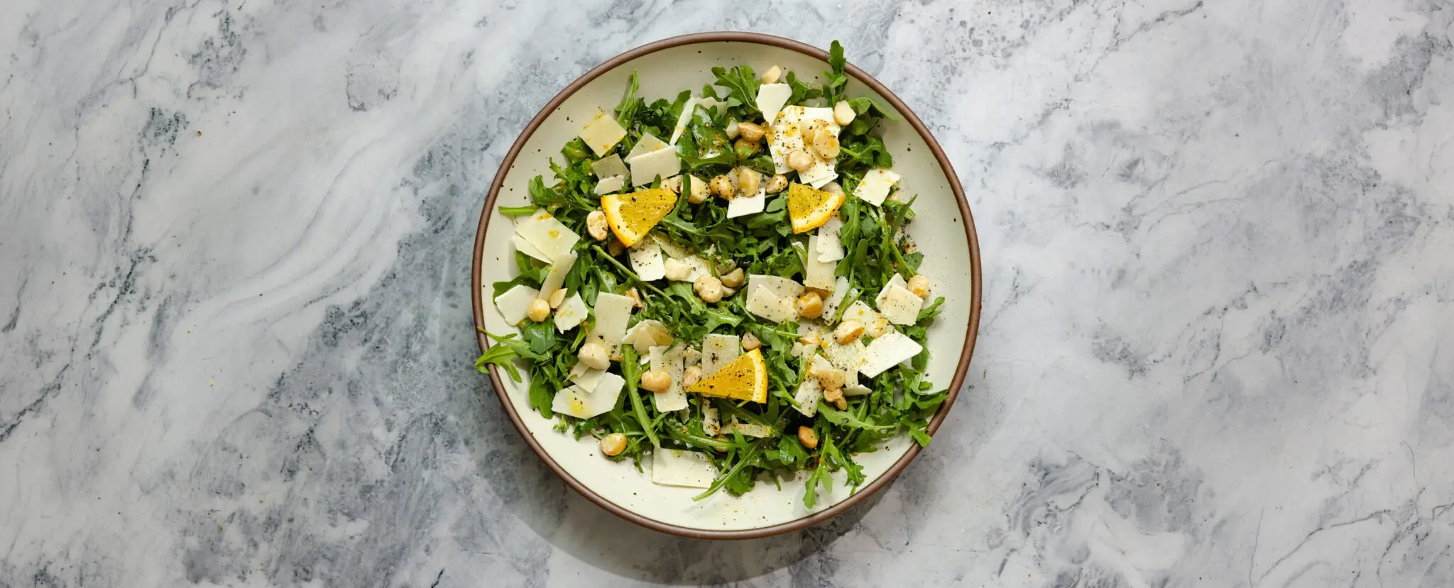 Overhead view of a Citrus Arugula Salad in a brown-rimmed ceramic plate, garnished with shaved parmesan, lemon slices, and chopped macadamia nuts.