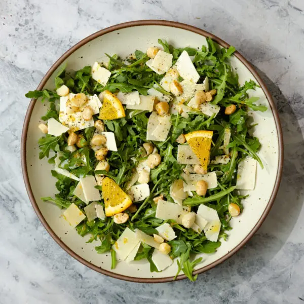 Overhead view of a Citrus Arugula Salad in a brown-rimmed ceramic plate, garnished with shaved parmesan, lemon slices, and chopped macadamia nuts.