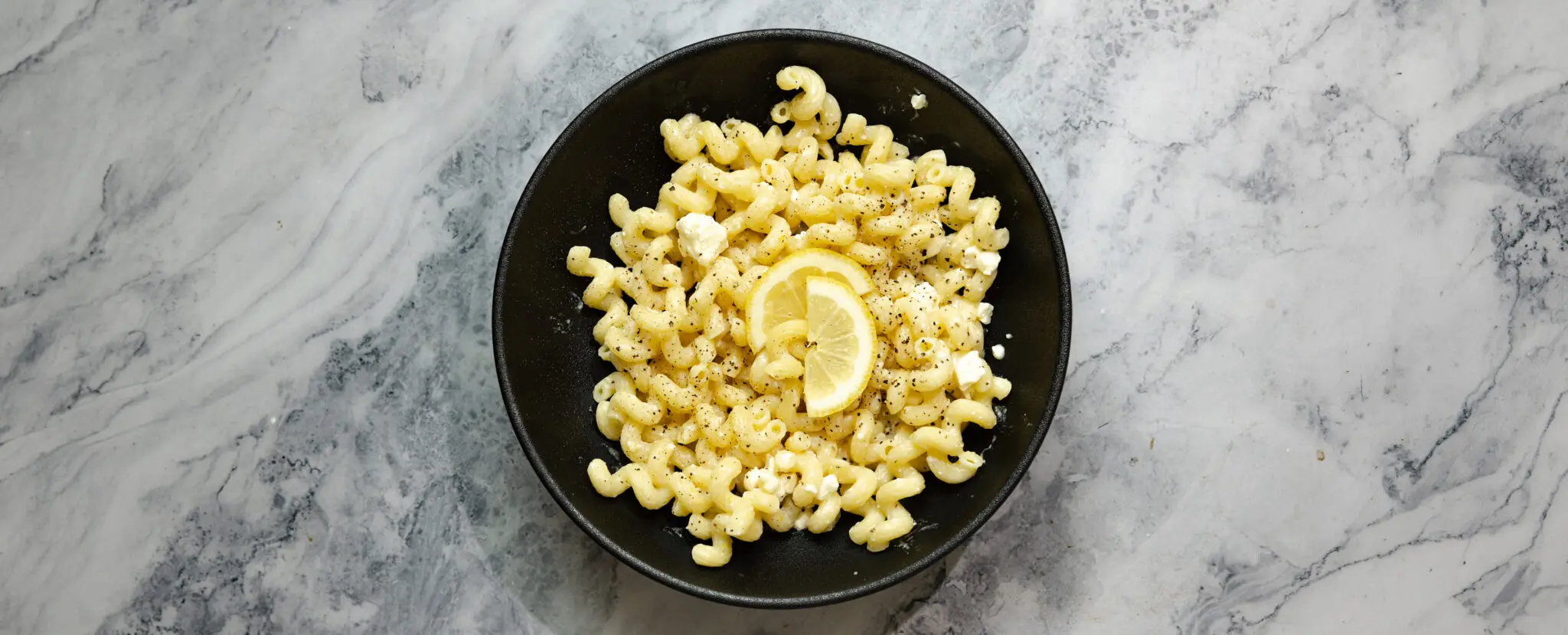 Overhead view of Lemon Feta Pasta in a round black bowl and garnished with a couple slices of lemon.
