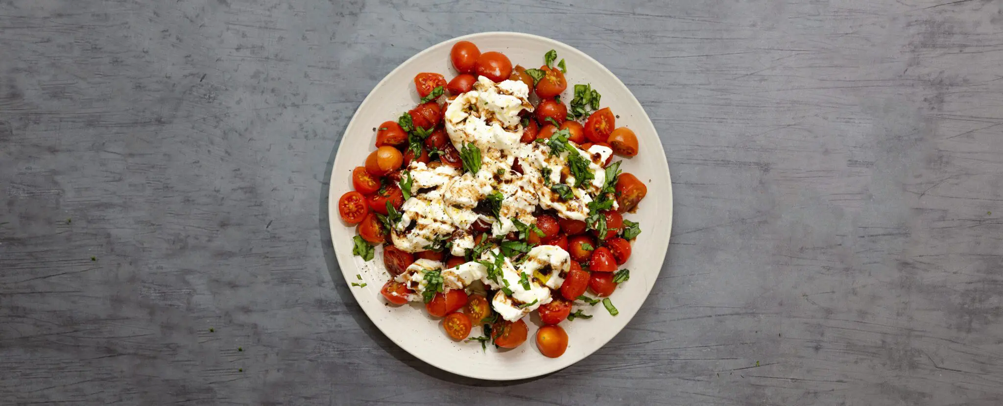 Overhead view of a white plate containing Burrata Caprese, served over a gray wood tabletop.