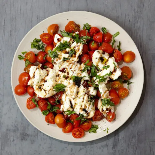 Overhead view of a white plate containing Burrata Caprese, served over a gray wood tabletop.