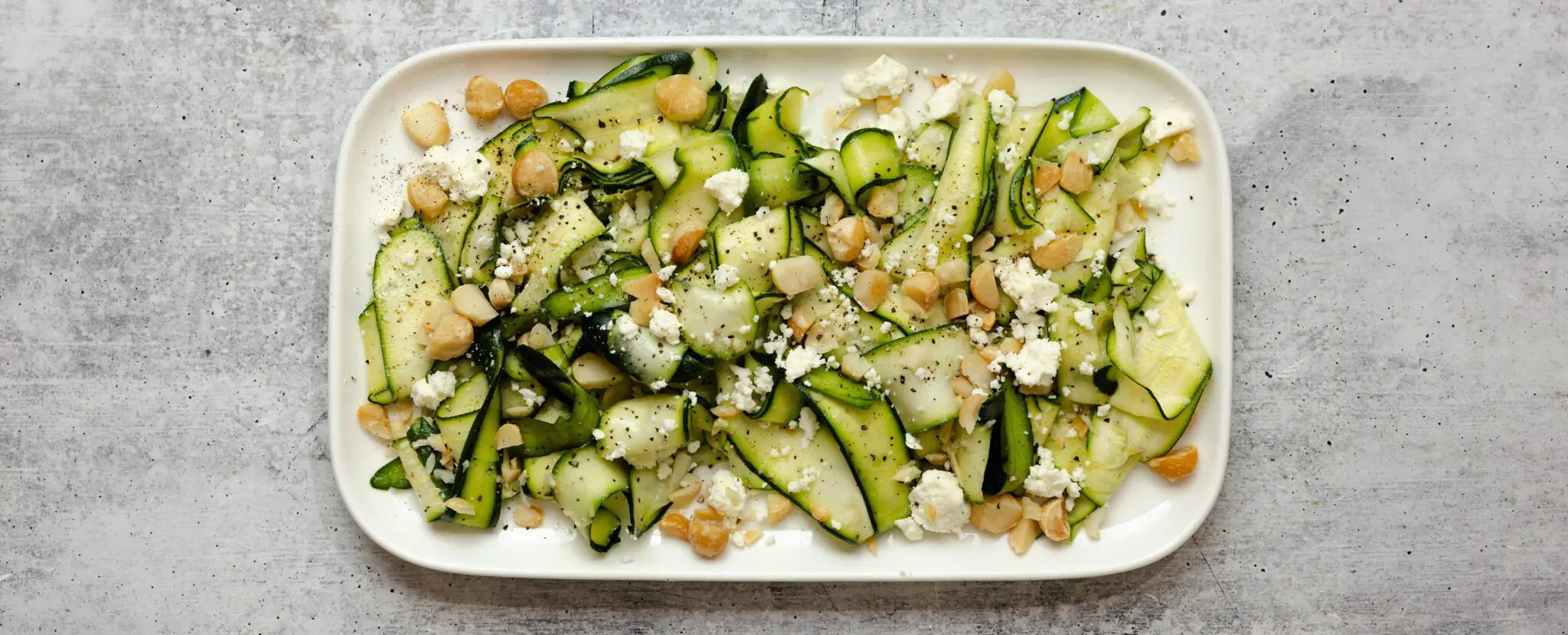 Overhead view of a Shaved Zucchini Salad garnished with crumbled feta and nuts, on a white large rectangular platter over a textured stone tabletop.