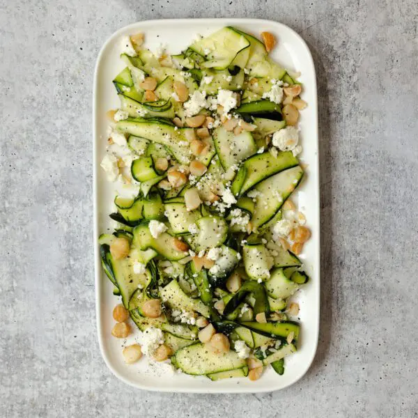 Overhead view of a Shaved Zucchini Salad garnished with crumbled feta and nuts, on a white large rectangular platter over a textured stone tabletop.