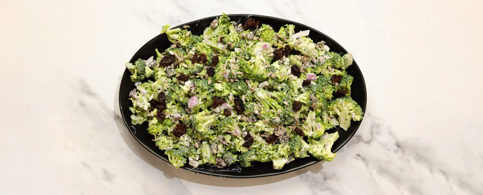 Overhead view of a large oval plate with Creamy Crunchy Broccoli Salad, served over a white marble tabletop.