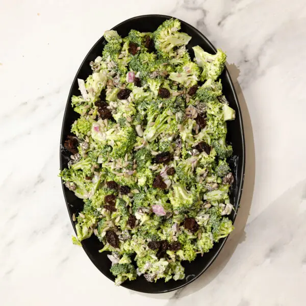 Overhead view of a large oval plate with Creamy Crunchy Broccoli Salad, served over a white marble tabletop.