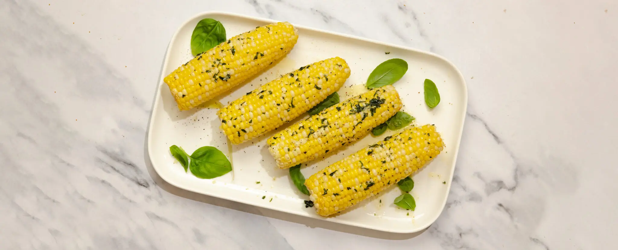 Overhead view of a large rectangular plate holding four cobs of herby basil butter corn garnished with basil leaves, served over a white marble tabletop.