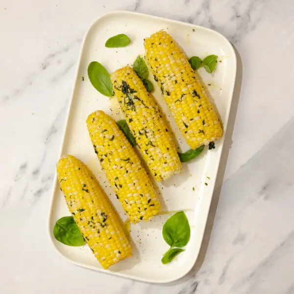 Overhead view of a large rectangular plate holding four cobs of herby basil butter corn garnished with basil leaves, served over a white marble tabletop.