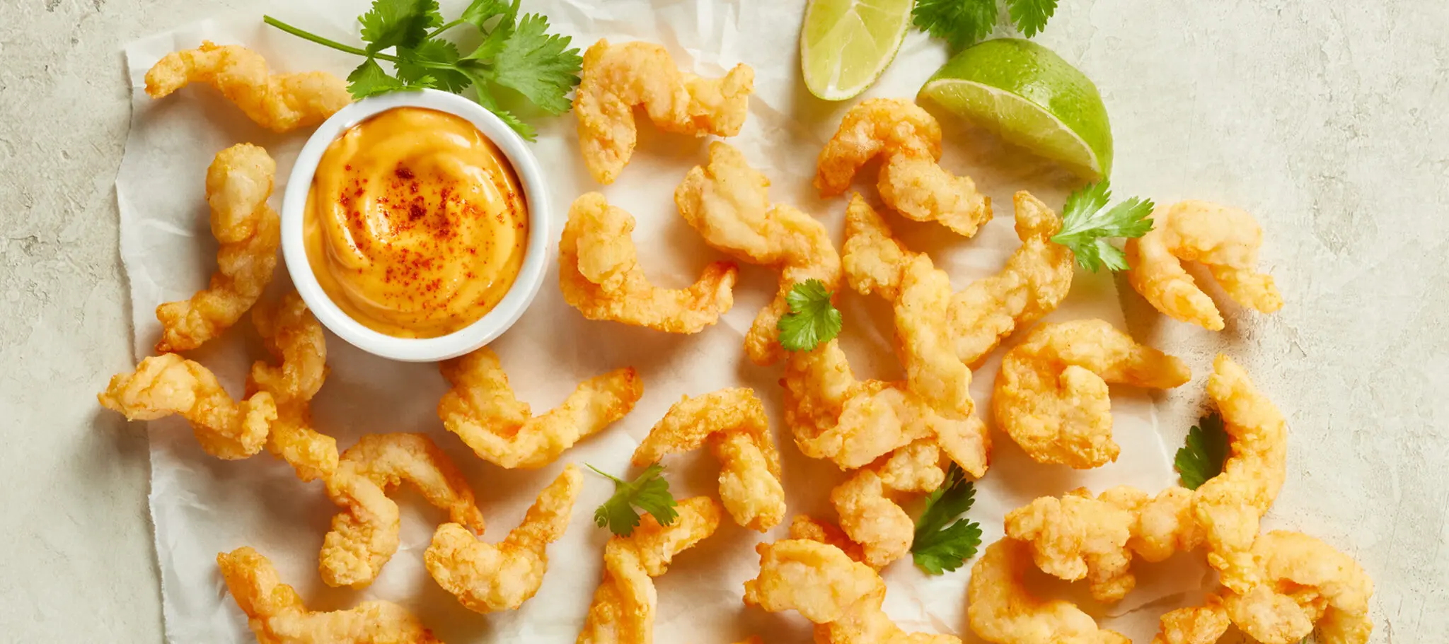 Social Kitchens Professional Corkscrew Shrimp pieces on parchment paper with lime wedges, some parsley, and a side dish of aioli sauce.