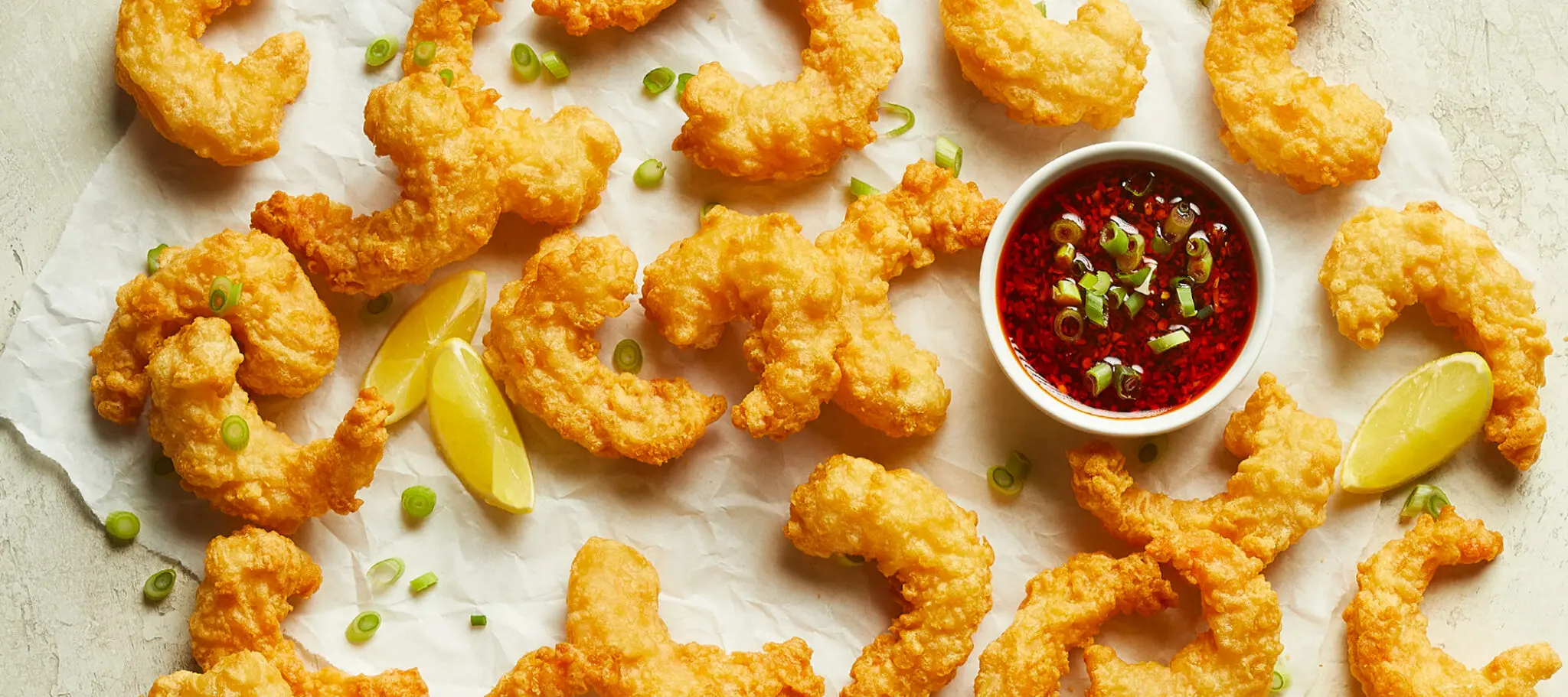 Social Kitchens Professional Crispy Battered Shrimp pieces on parchment paper with lemon wedges, some green onion, and a side dish of sauce.