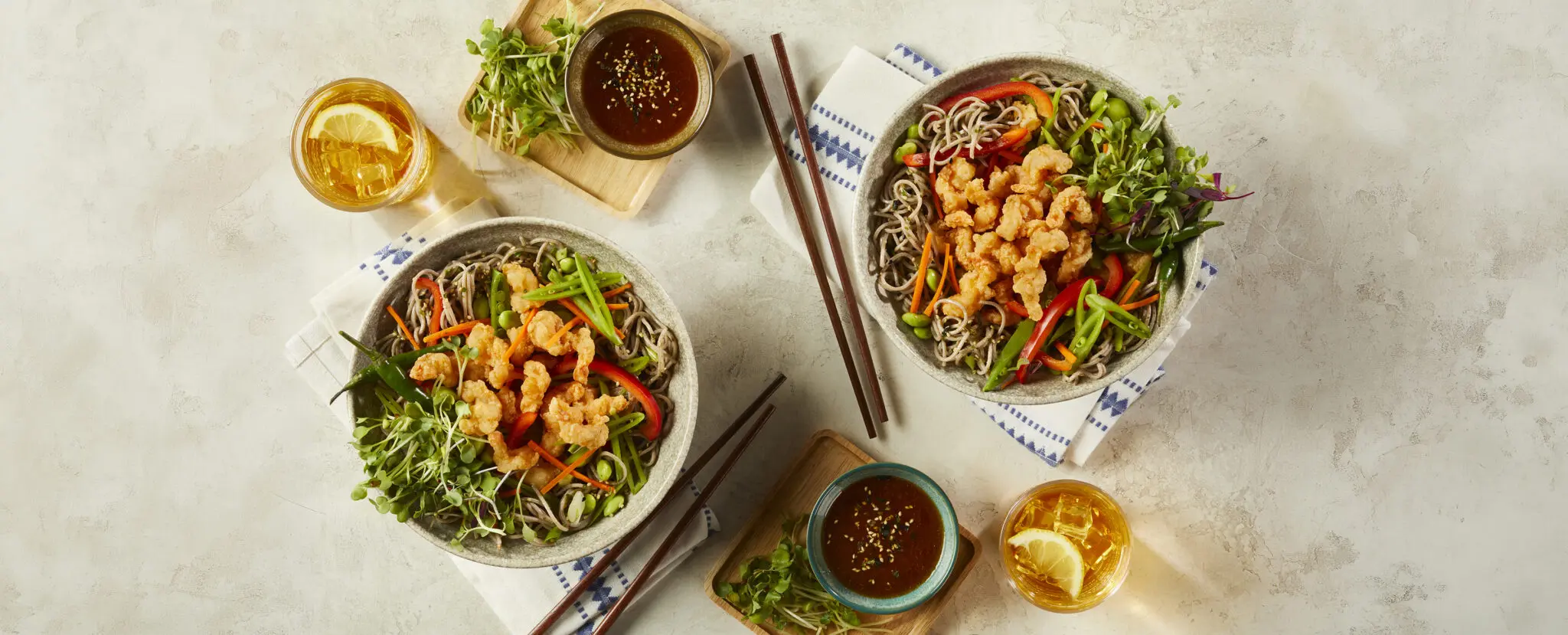 Overhead view of two Cold Soba Noodle Shrimp Salad bowls served with additional sauces on the side and two lemon iced teas, over a beige stone table surface.