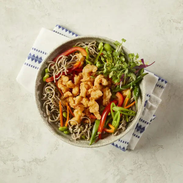 Overhead view of a bowl of Cold Soba Noodle Shrimp Salad served over a white and blue striped towel and beige stone table surface.