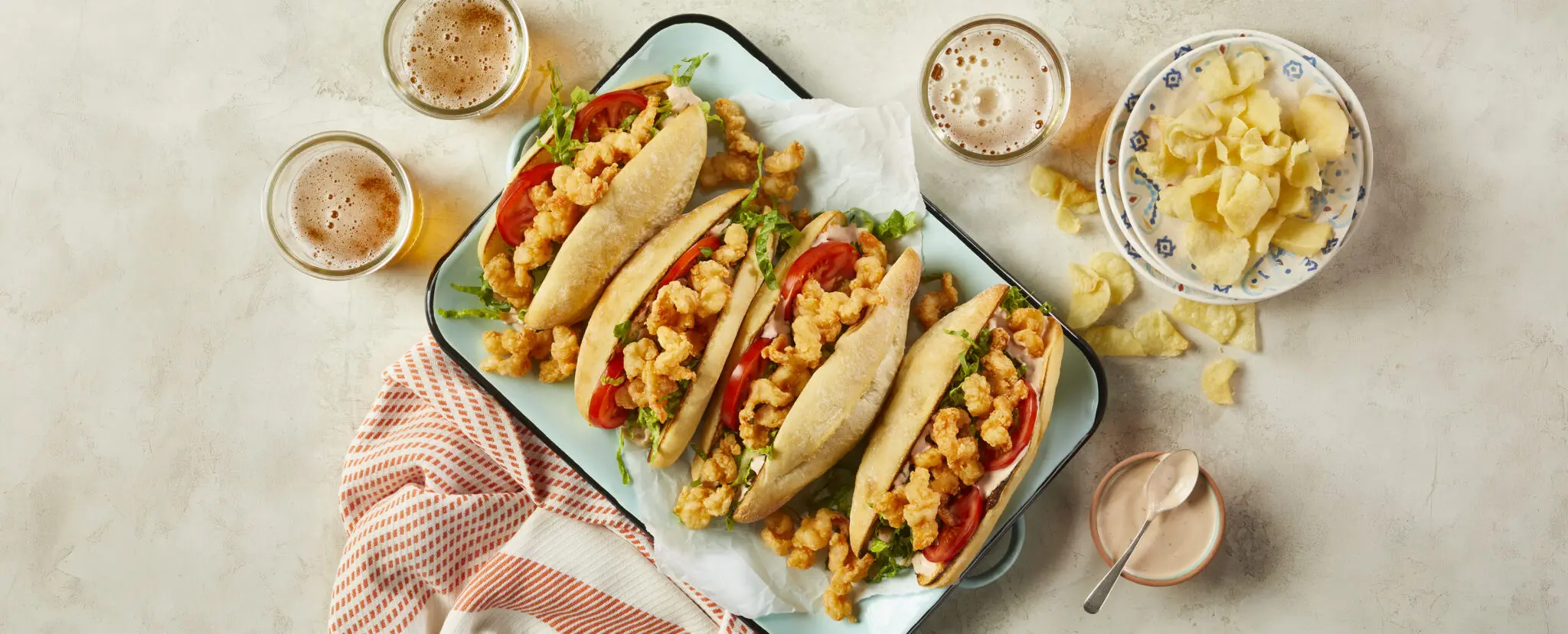 Overhead view of four Shrimp Po' Boy sandwiches lined up on a rectangular dish alongside a plate of potato chips, a side of aioli sauce, and three glasses of beer.
