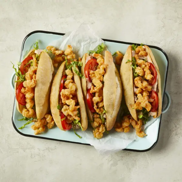 Overhead view of four Shrimp Po' Boy sandwiches lined up on parchment paper within a rectangular dish over a beige stone table surface.