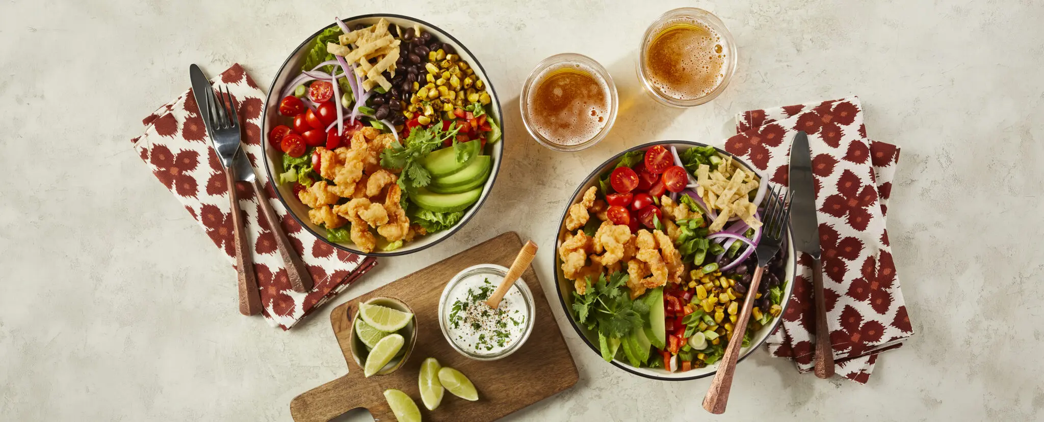 Overhead view of two Corkscrew Shrimp Southwest Salad bowls using Social Kitchens Professional Corkscrew Shrimp. To complete the table setting are cutlery, patterned cloth napkins, and two glasses of iced lemon tea.