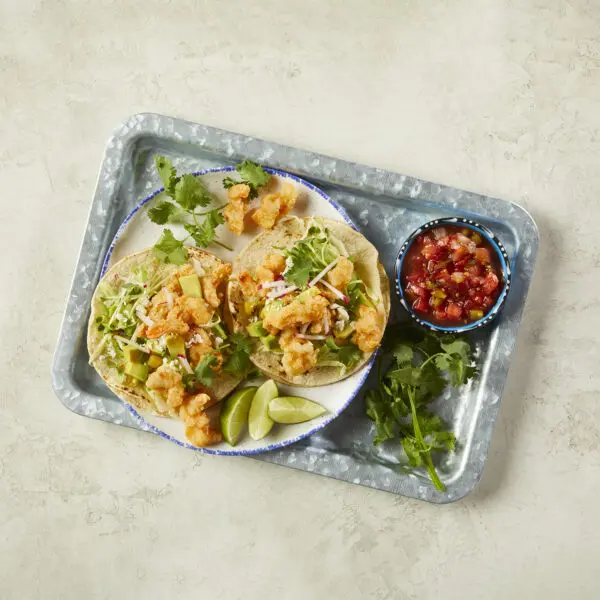 Overhead view of four Corkscrew Shrimp Tacos made with Social Kitchens Professional Corkscrew Shrimp. Garnished with microgreens, lime wedges, cilantro, and cotija cheese.