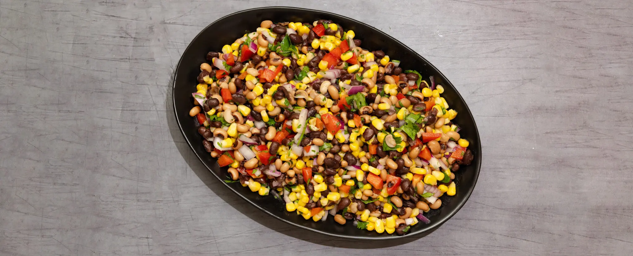 Overhead view of a large oval black plate of Cowboy Caviar which consists of corn, black-eyed peas, red bell peppers, cilantro, and red onion, served over a gray table surface.
