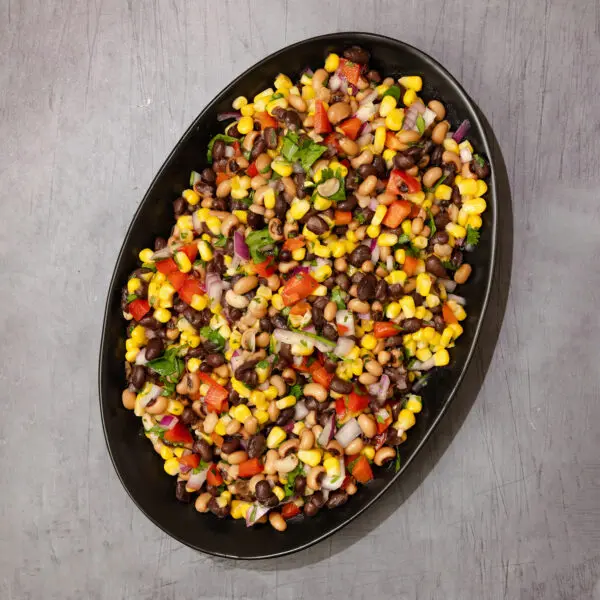 Overhead view of a large oval black plate of Cowboy Caviar which consists of corn, black-eyed peas, red bell peppers, cilantro, and red onion, served over a gray table surface.