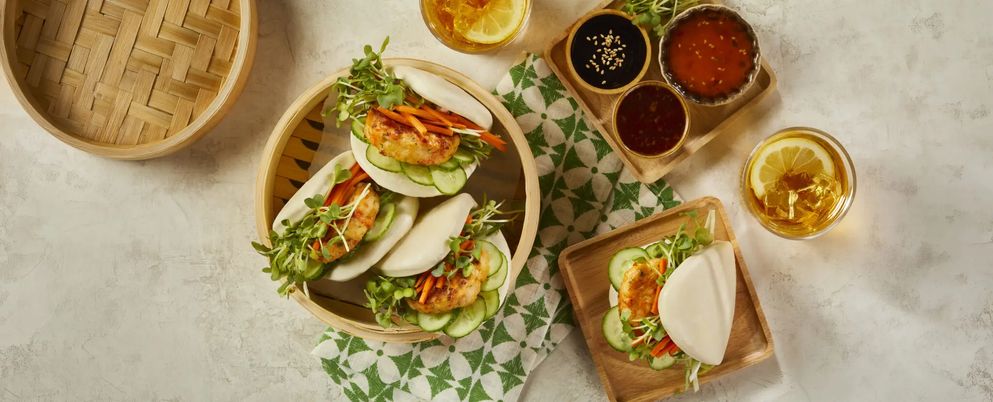Overhead view of three Shrimp Bao buns using Social Kitchens Premium Shrimp Sliders microgreens, carrots, and cucumbers in a pillow soft bao bun. Served in a traditional wooden steam basket over a stone table surface. Off to the side is a single Shrimp Bao Bun on a square plate, a small tray of assorted dipping sauces, and a glass of iced lemon tea.
