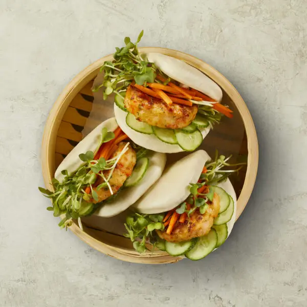 Overhead view of three Shrimp Bao buns using Social Kitchens Premium Shrimp Sliders microgreens, carrots, and cucumbers in a pillow soft bao bun. Served in a traditional wooden steam basket over a stone table surface.
