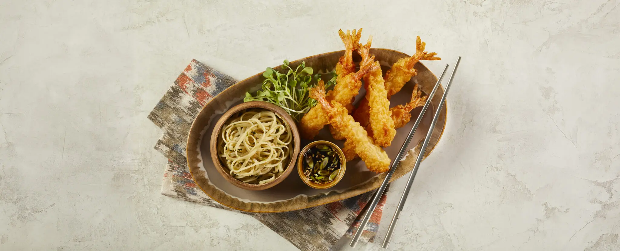 Overhead view of Social Kitchens Professional Tempura Battered Shrimp in an oval bowl with a small round bowl of noodles, another smaller bowl of soy sauce with green onions and sesame seeds, and a small handful of microgreens. This Tempura Shrimp Soy Garlic Noodles dish is served over a pattern-printed cloth napkin over a beige stone table surface.