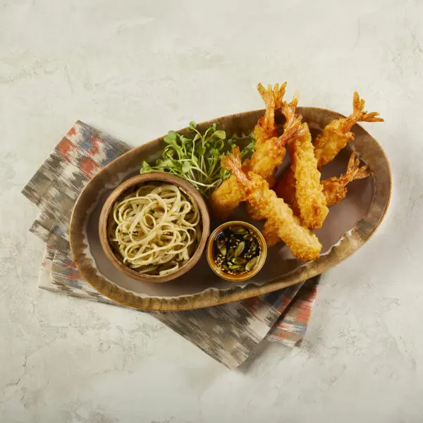 Overhead view of Social Kitchens Professional Tempura Battered Shrimp in an oval bowl with a small round bowl of noodles, another smaller bowl of soy sauce with green onions and sesame seeds, and a small handful of microgreens. This Tempura Shrimp Soy Garlic Noodles dish is served over a pattern-printed cloth napkin over a beige stone table surface.