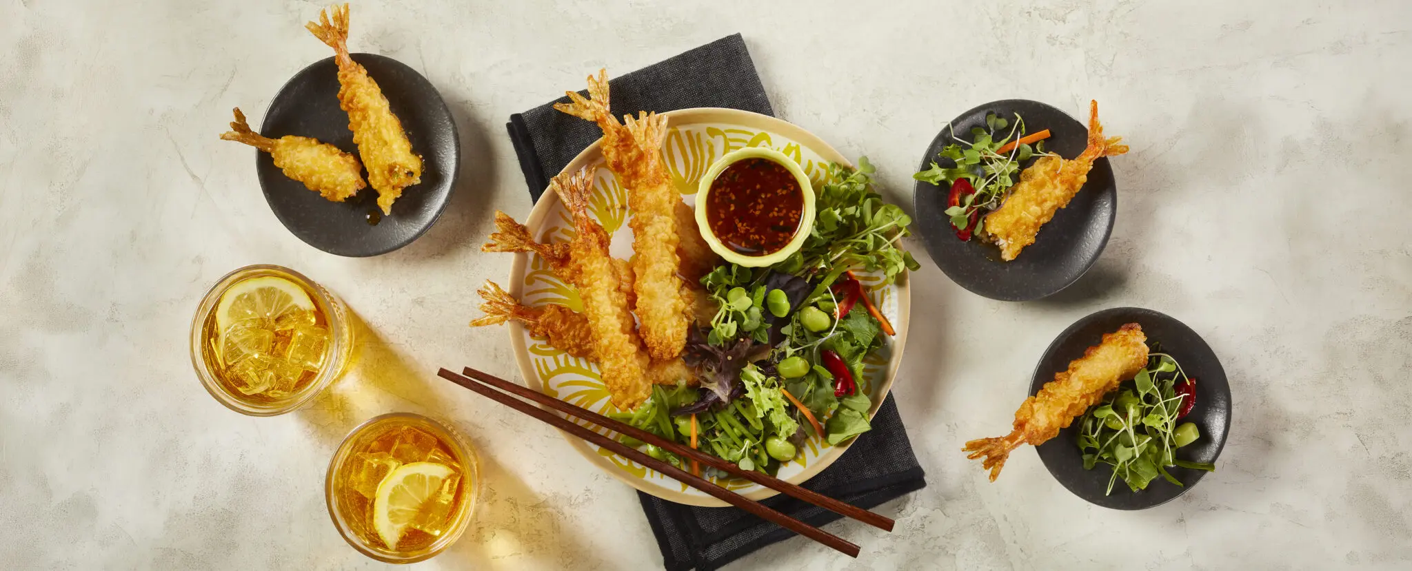 Overhead view of a bowl of Social Kitchens Professional Tempura Battered Shrimp with a side salad and chili ginger dressing in a tiny ramekin. This shrimp tempura with side salad is served with a pair of chopsticks resting over the bowl and a black cloth napkin underneath, over a beige stone table surface. Surrounding the center bowl are two glasses of lemon iced tea and three smaller bowls each with a piece of shrimp and salad.