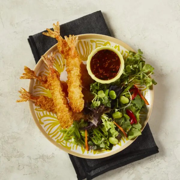 Overhead view of a bowl of Social Kitchens Professional Tempura Battered Shrimp with a side salad and chili ginger dressing in a tiny ramekin. This shrimp tempura with side salad is served over a black cloth napkin over a beige stone table surface.