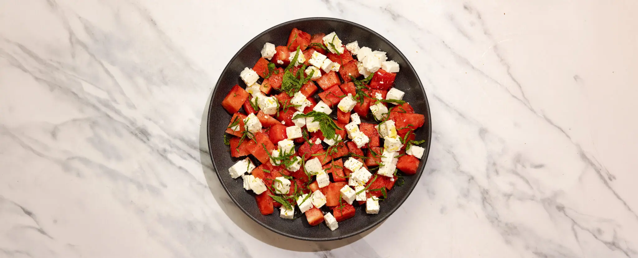 Overhead view of Watermelon Feta Mint Salad on a round black plate over a marble table surface.