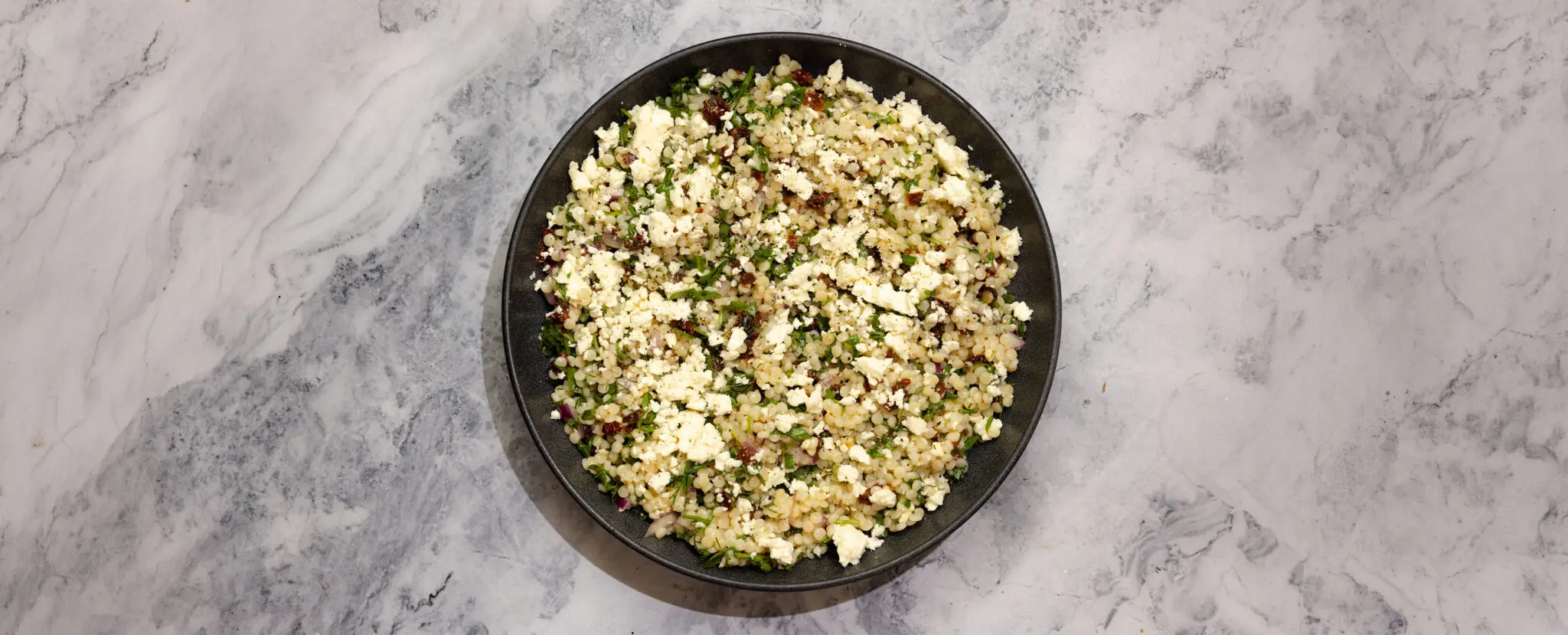 Overhead view of couscous pasta salad on a black plate over a marble table top.