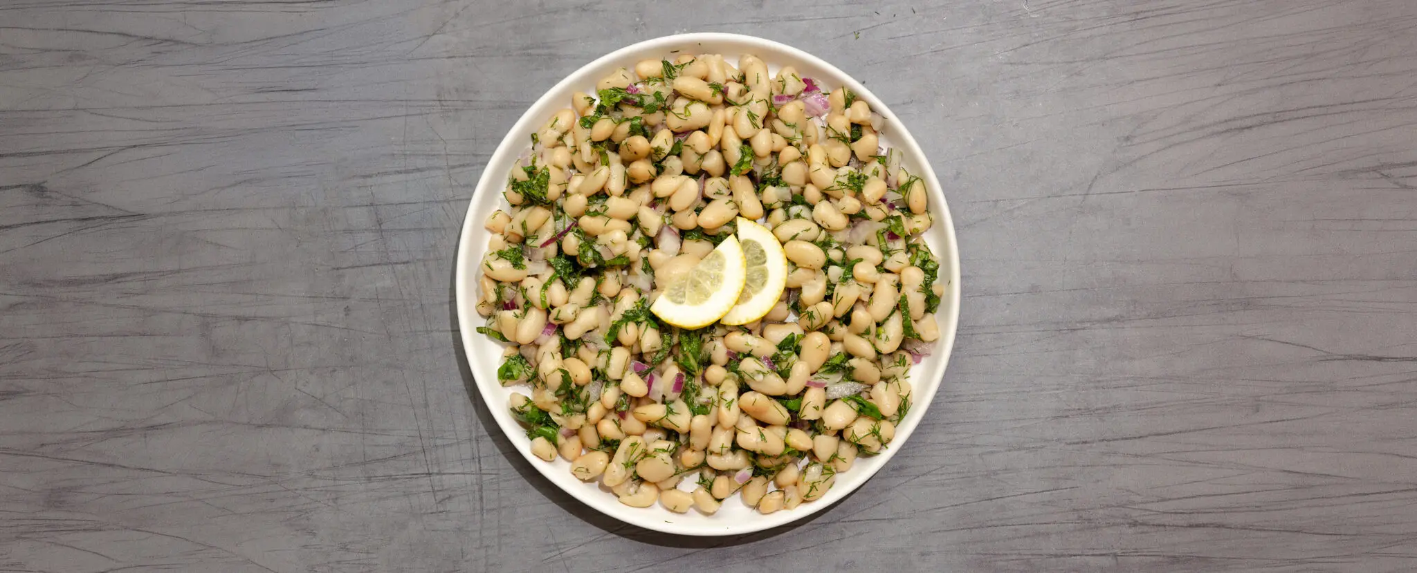 Overhead view of an Herby White Bean Salad in a white bowl garnished with two half slices of lemon and served over a gray wood table top.